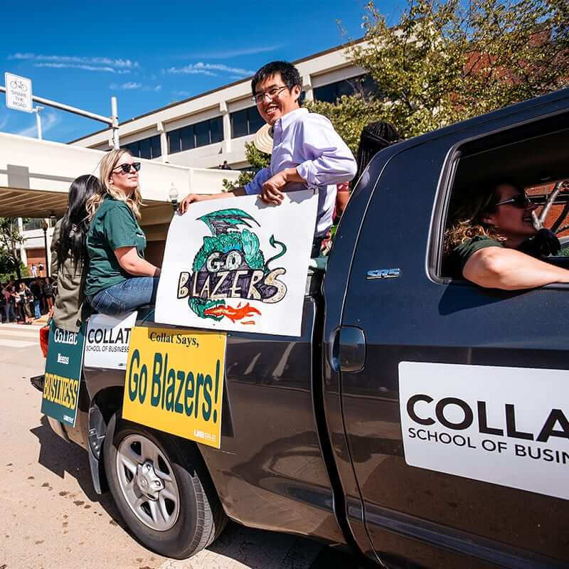 Students riding in the back of a pickup truck at the homecoming parade, smiling and waving at the camera.