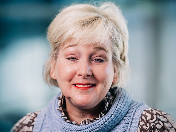 Headshot of a woman with light blond hair smiling, wearing a floral shirt and purple vest
