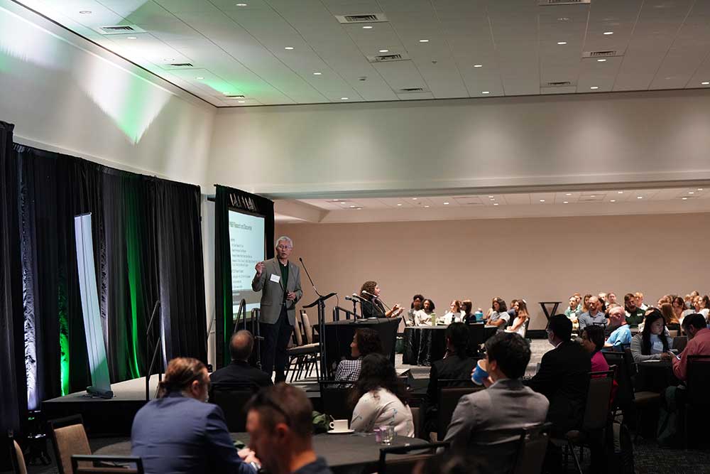 Dr. Vu Nguyen leading a presentation on a stage with screens behind him. He is presenting in front of a large audience of people all seated at tables.