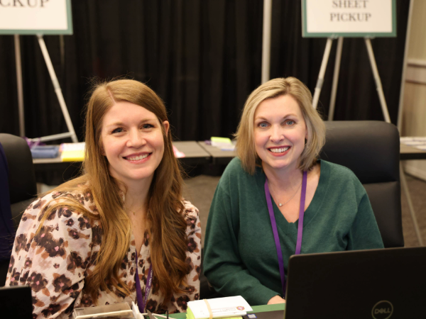 Two women in professional attire smiling for a photo at a registration desk at a conference