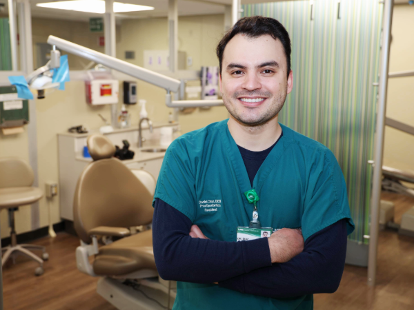 A dental resident in green scrubs stands in a clinical operatory with arms crossed, smiling at the camera. A dental chair and overhead equipment are visible behind him in a treatment area.