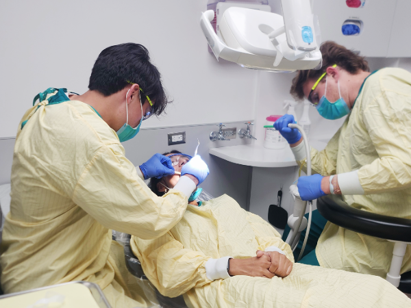 Two dental students in masks, gloves, gowns, and protective eyewear perform mock treatment on a reclining student—one uses a lighted instrument near the mouth while the other operates suction; the student lies back with hands clasped.