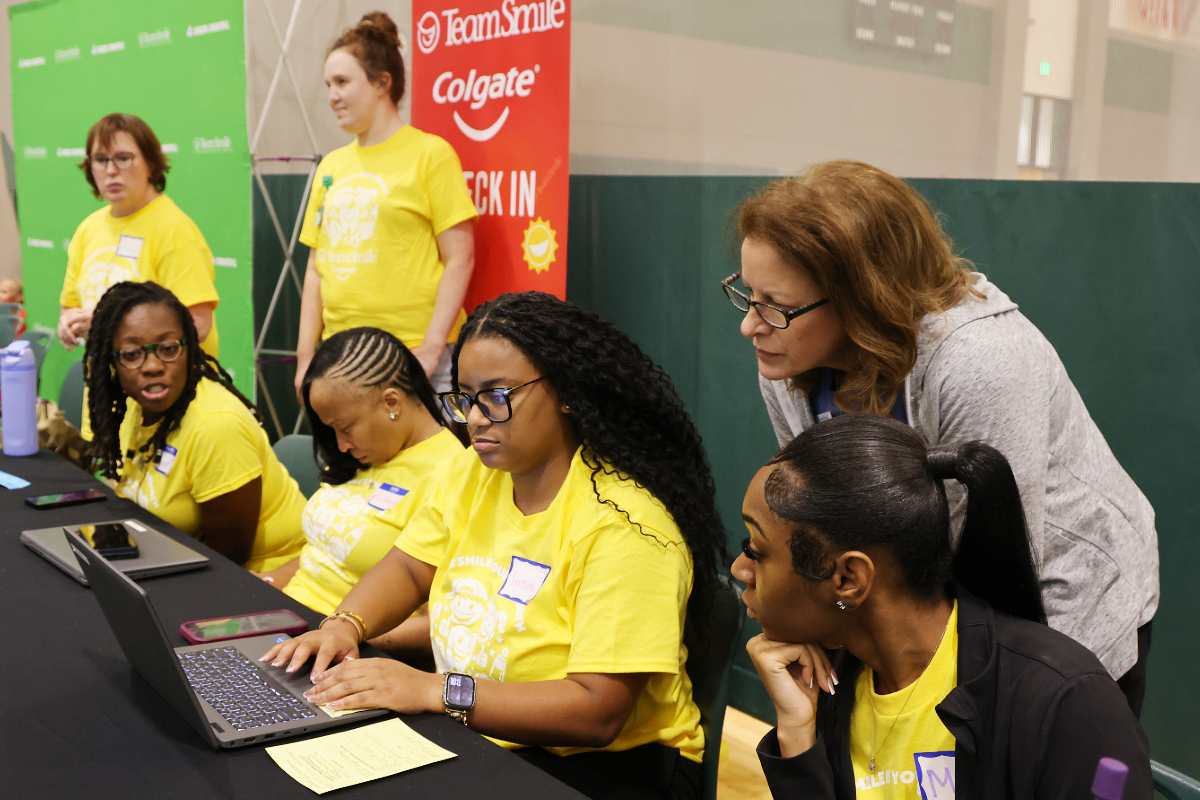 Volunteers staffing a TeamSmile check-in table: women in bright yellow event shirts working on laptops and forms while another staff member leans over to assist.