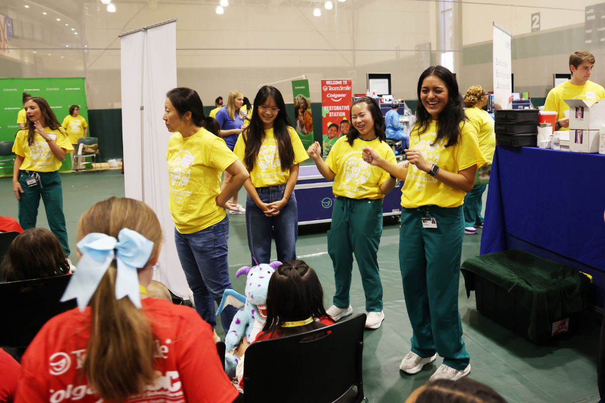 Dental students and volunteers in yellow TeamSmile shirts smile and perform an upbeat dance at dental outreach event for seated children, one with a plush toy.