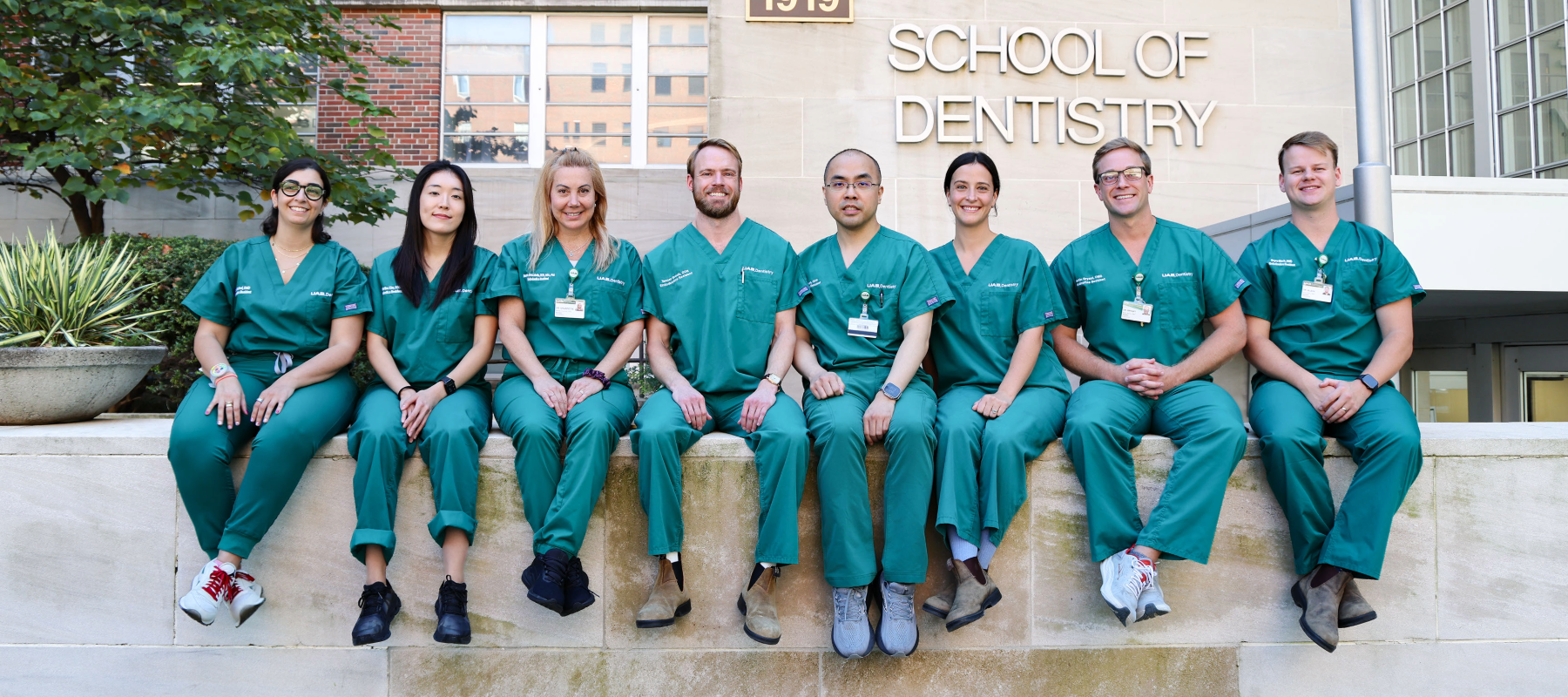 Group photo of endodontics residents in green scrubs sitting on concrete wall in front of the UAB School of Dentistry building