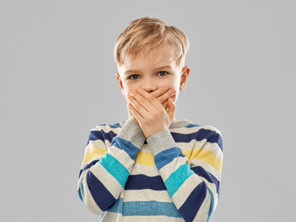 shocked little boy in striped pullover closing his mouth by hands over grey background