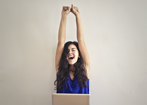 A young woman seated in front of her laptop excitedly raises her hands in the air.