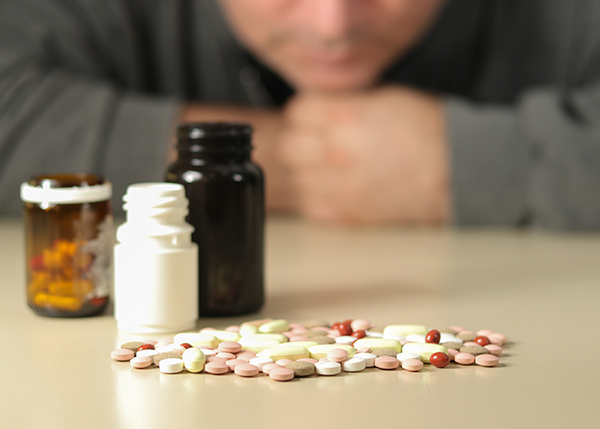 the bottom half of a man's face can be seen in the background looking at a table full of prescription bottles and pills