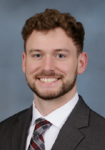 Dr. Wilson Fisher Professional headshot of a man wearing a dark suit jacket with a white shirt and a grey and red striped tie in front of a blue backdrop