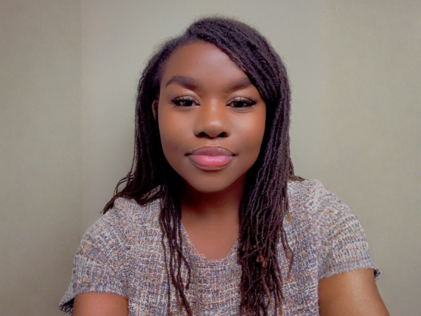 Headshot of a person with long hair, facing the camera against a plain light-colored background. The person is wearing a short-sleeved textured knit top and is centered in the frame with even indoor lighting.