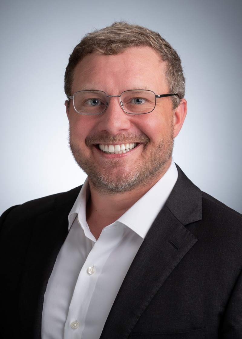 Photo of Tim Johnson, MD, smiling and wearing glasses, a black blazer, and white buttoned shirt against a soft grey background.