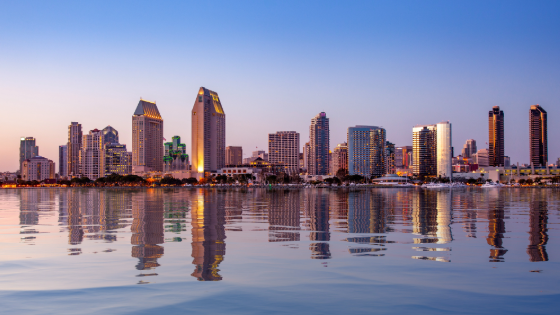 Skyline of downtown San Diego at dusk, with high-rise buildings illuminated against a blue and pink evening sky. The city lights reflect across calm water in the foreground, creating mirrored vertical streaks on the surface.