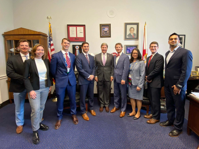 Nine people stand together for a meeting photo in an office setting, surrounded by official furnishings, flags, and framed displays.