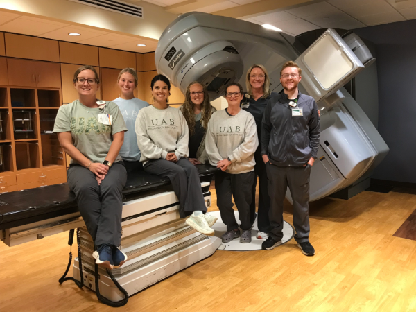 Group photo of seven people posing in a radiation oncology treatment room. Three people sit on the treatment table while four stand beside it, in front of a large gray linear accelerator used for cancer treatment.