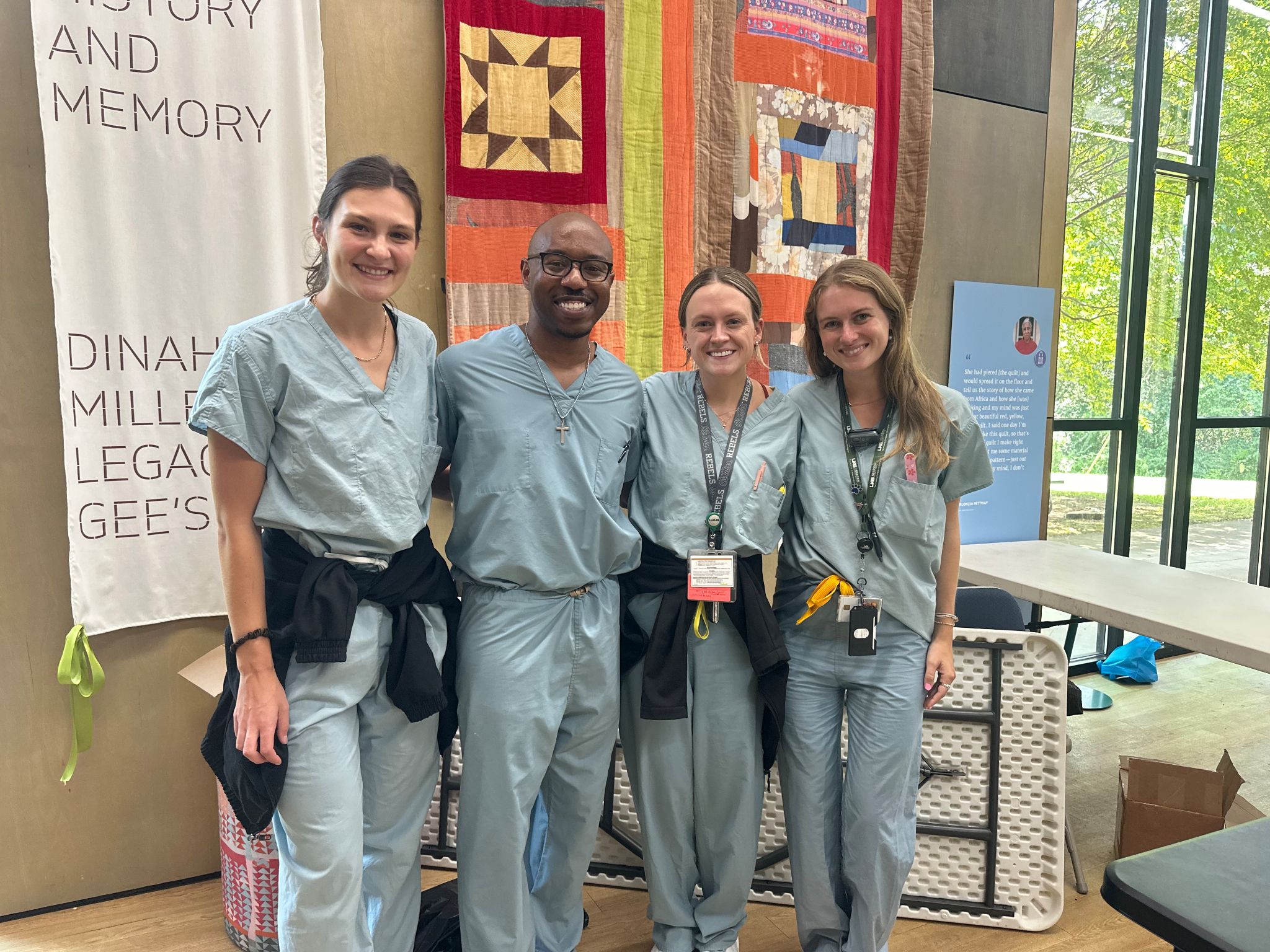 Four medical trainees in light blue scrubs stand together indoors in front of a colorful quilt display. Several ID badges and lanyards are visible, and a banner reading “History and Memory” with references to Gee’s Bend hangs beside the exhibit. Folding tables and tall windows overlooking greenery are visible in the background.