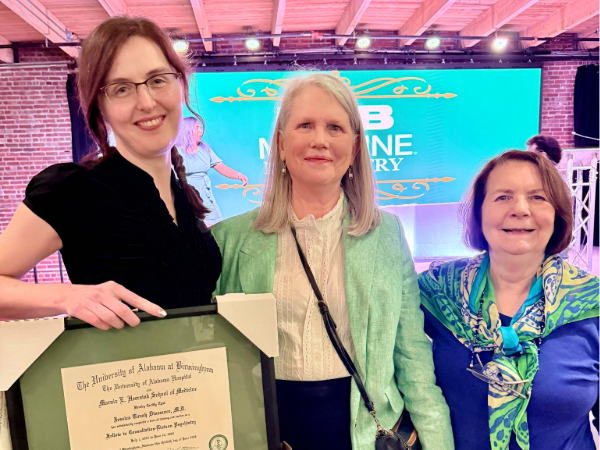 Three adults pose indoors holding a framed University of Alabama at Birmingham certificate in front of a lit stage.
