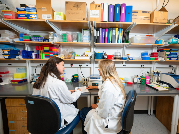 Two women in white coats talk and work in a lab.