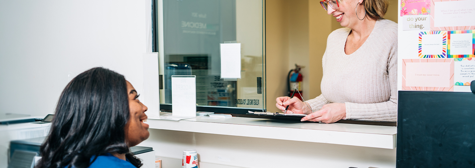 Woman at a reception window smiles while writing on a clipboard as a staff member in scrubs sits at a desk.