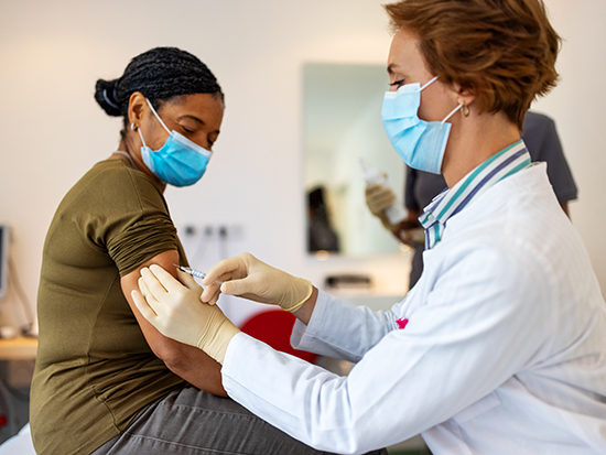 Female doctor wearing a face mask giving vaccine to a mature patient in her clinic. General practitioner giving flu shot to a senior woman in clinic.