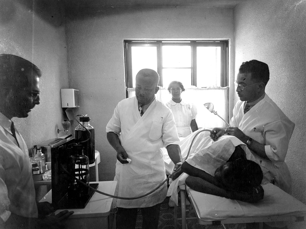 In a historic black-and-white photograph, doctors and a nurse treat a patient at Slossfield Community Center.