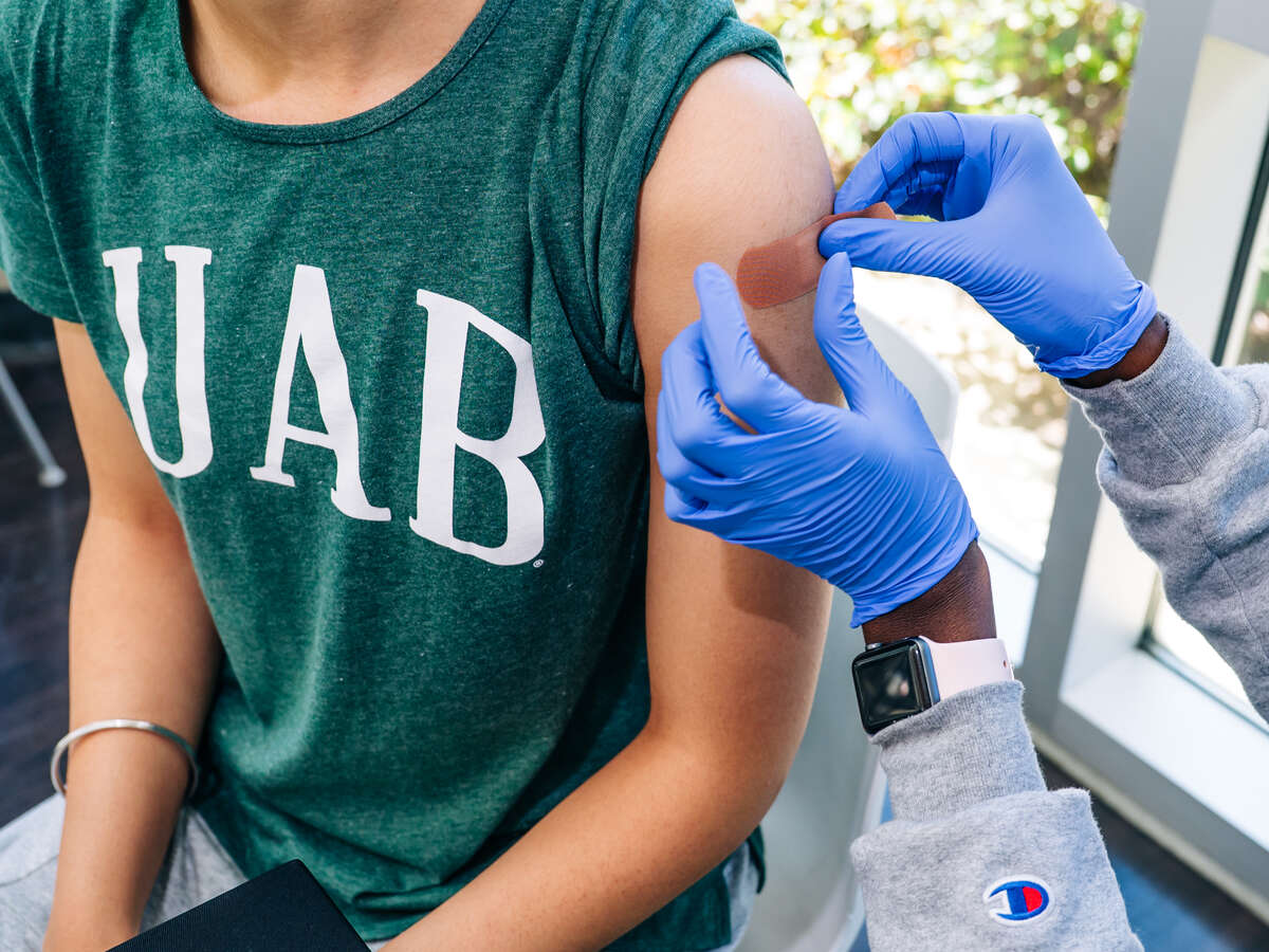 Flu Shot Stop, Hill Student Center. Close-up, hands of nursing student are wearing PPE gloves while applying a bandage over influenza injection site in the arm of a seated female student wearing a green UAB t-shirt, October 2025.