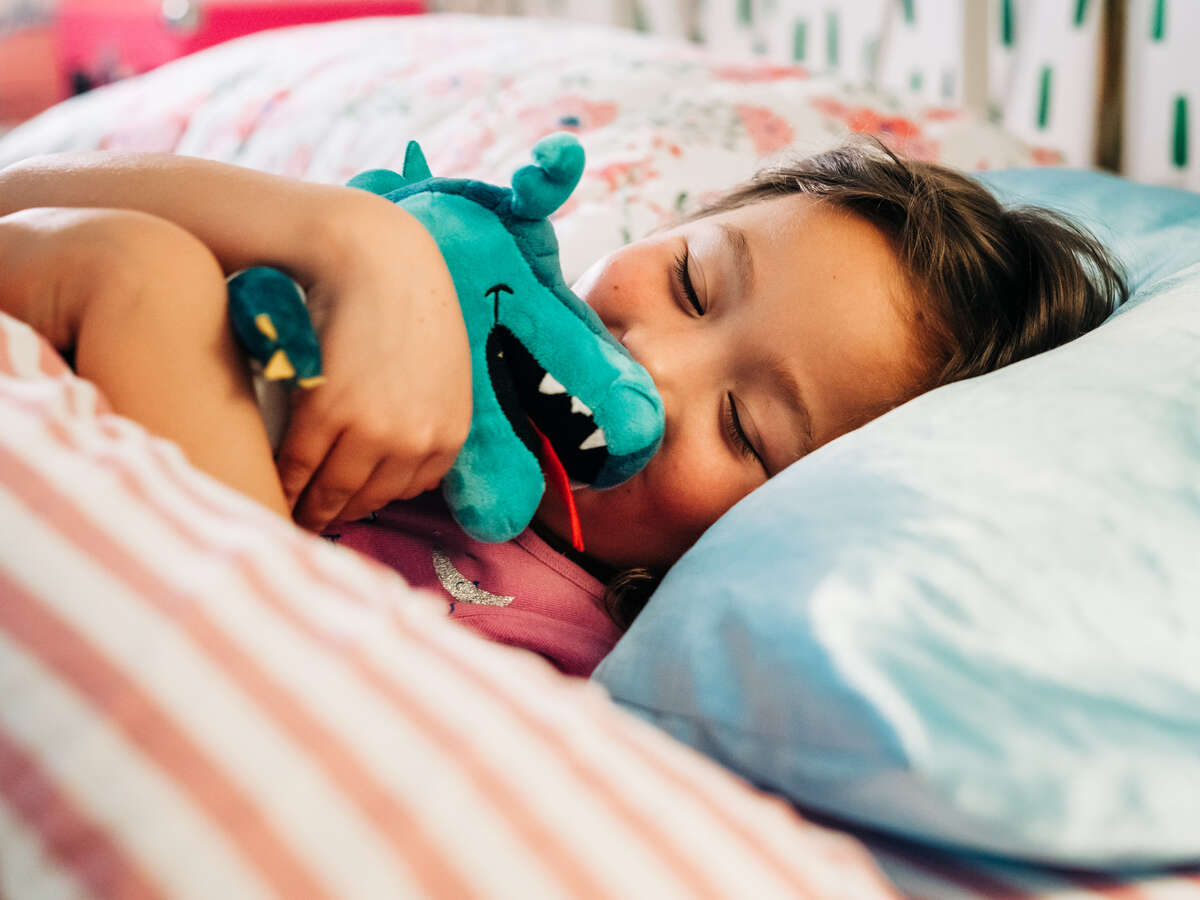 Close-up of smiling girl hugging a Blaze (mascot) plushie stuffed animal toy while waking up in bed after sleeping in home.