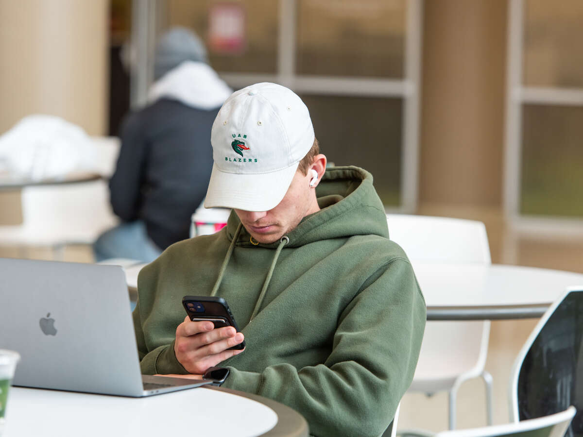 Male student is wearing a green sweatshirt and a "UAB" cap while working on a laptop computer and mobile phone while sitting at a table in the dining area of the Hill Student Center.