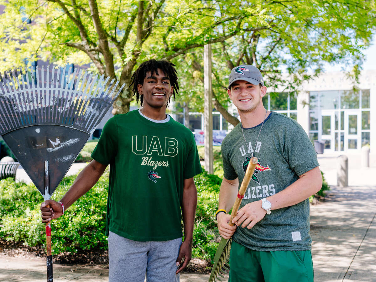 Two men representing UAB and wearing UAB clothing are holding rakes while helping to clean up the grounds of Carver High School as a community service project.
