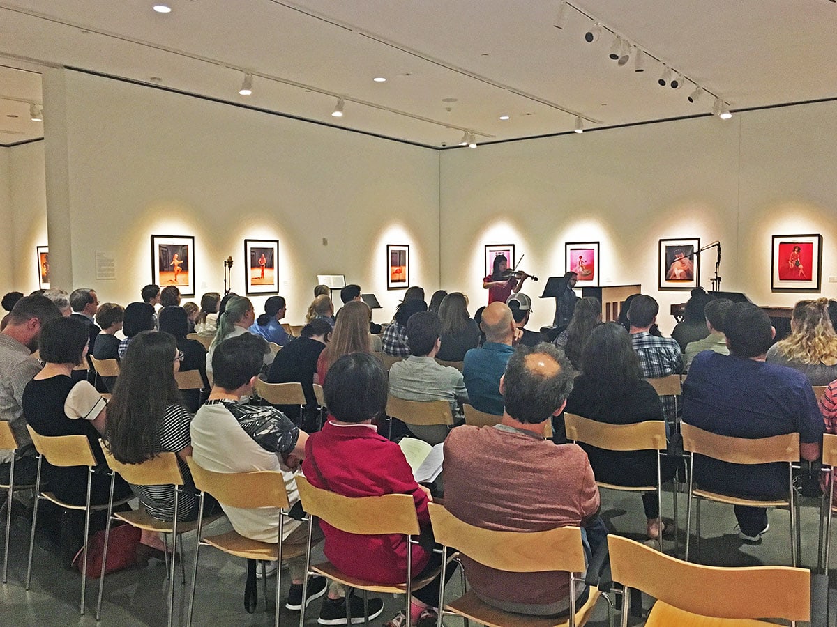 n audience sits inside a large art gallery with two musicians performing on violin and piano. Art illuminated by gallery lights hangs on the walls around them.