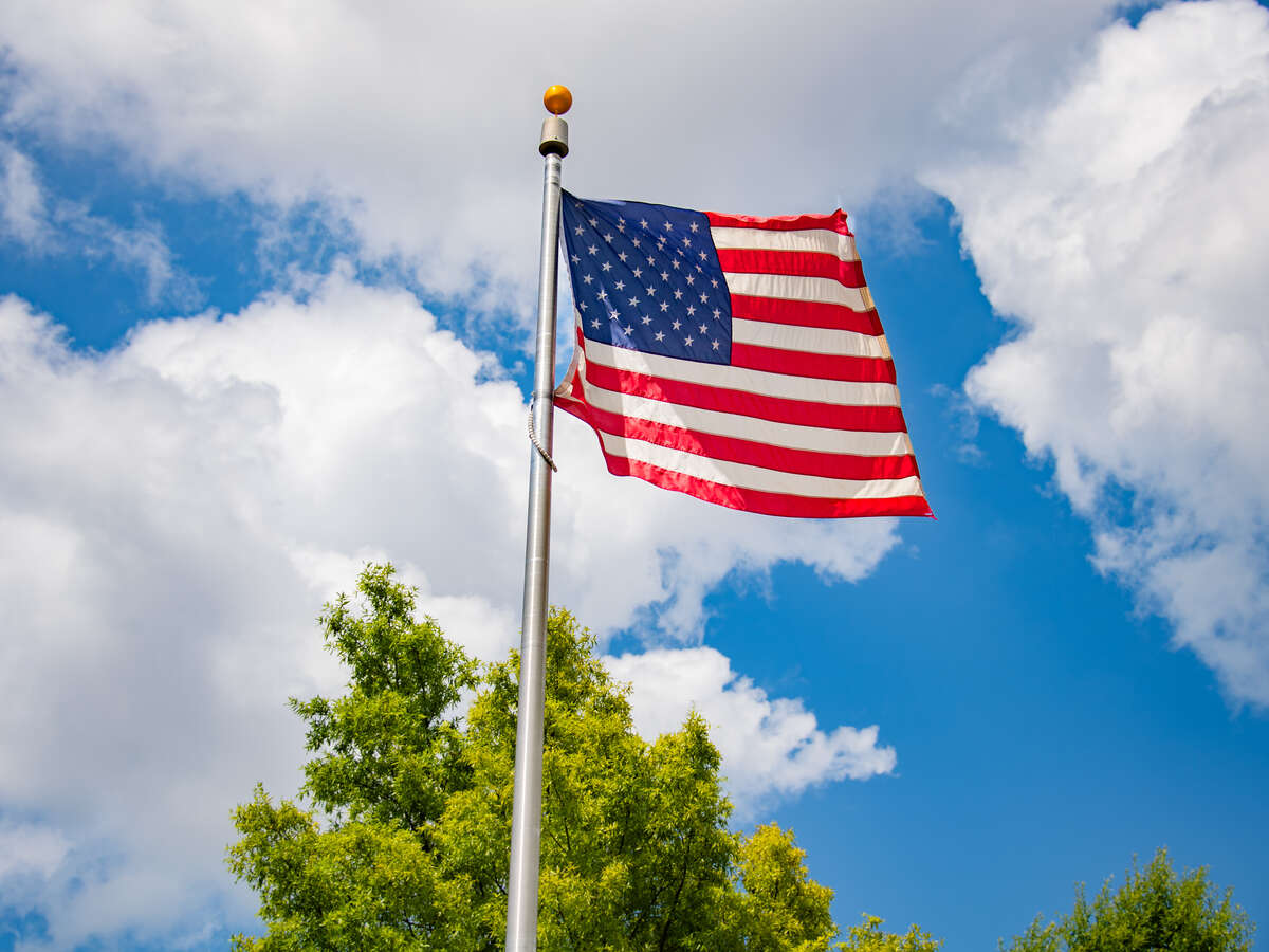 American flag flying on flagpole on the Campus Green with blue sky, white clouds, and the top of a green tree in the background.