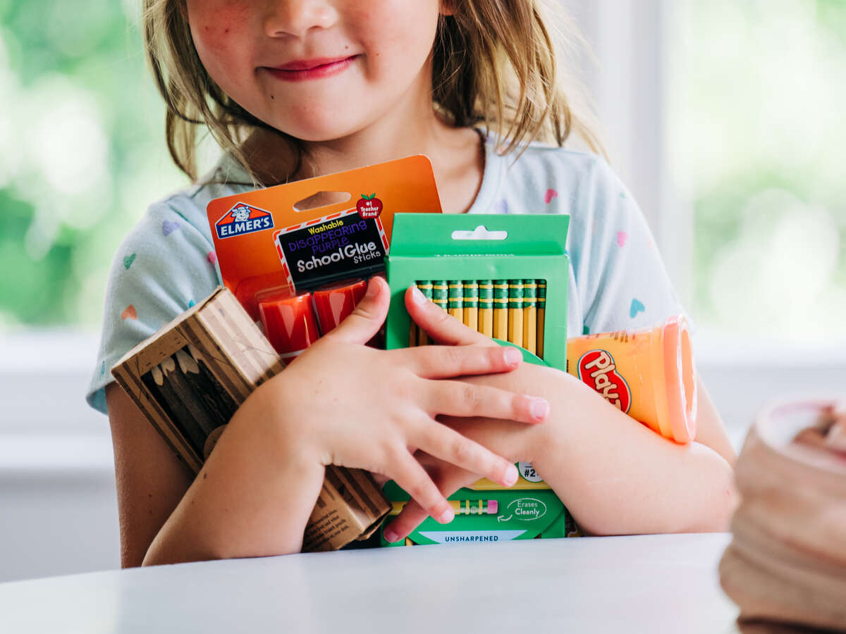 Close-up of smiling girl holding new school supplies, including pencils, crayons, and glue sticks, while standing beside dining table in home, July 2025.