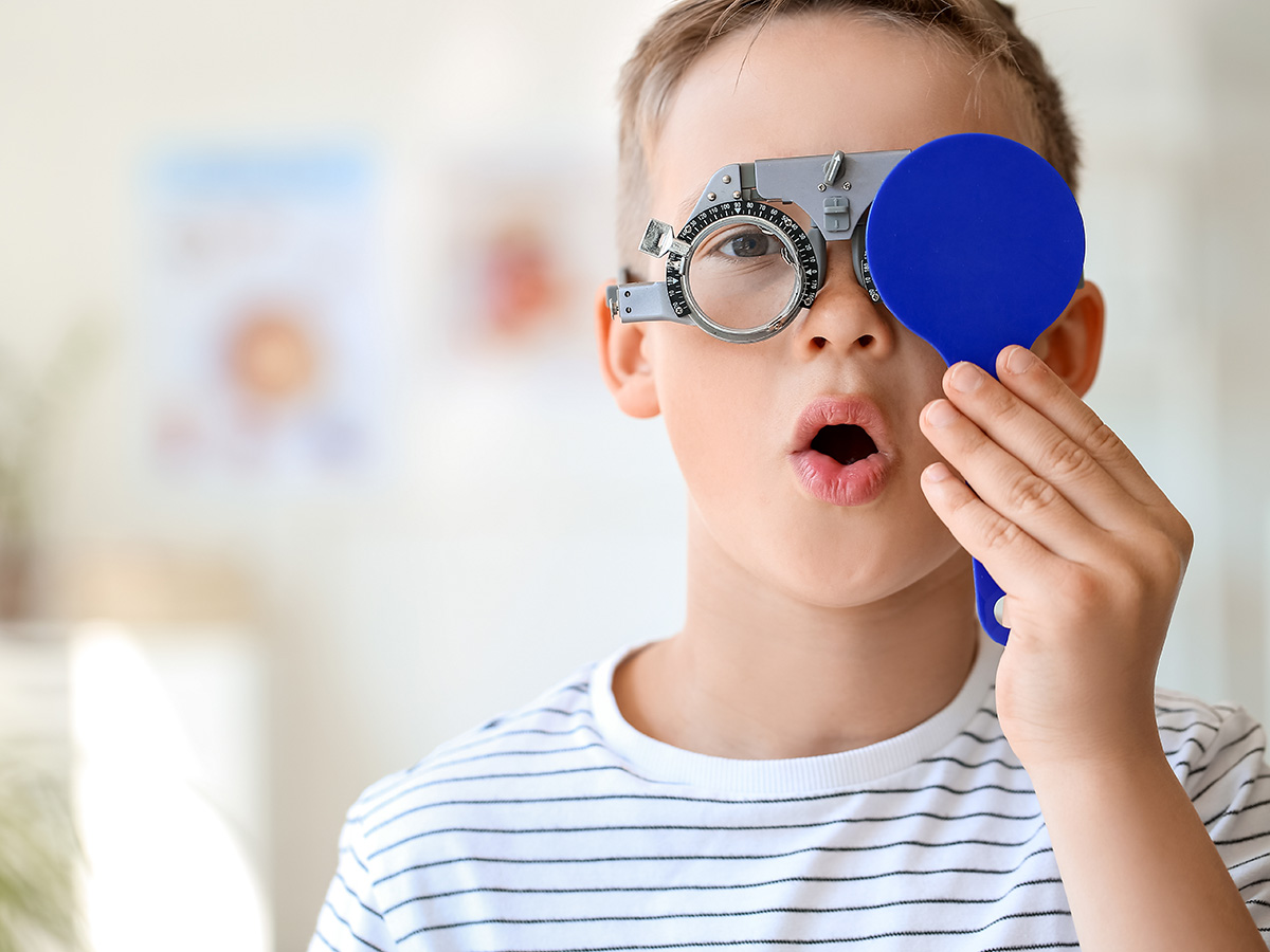 Boy in white shirt with blue stripes covers left eye with blue eye test tool during eye exam.