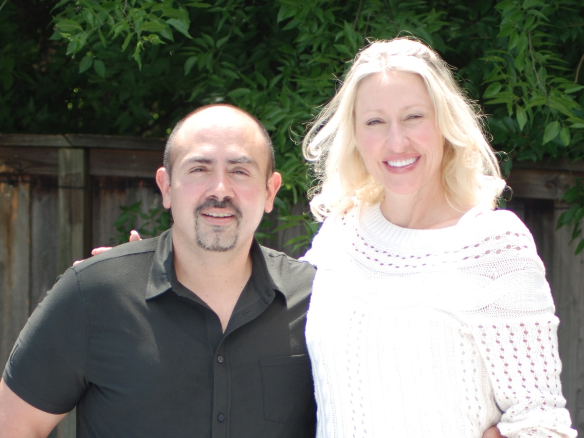 Outside photo of Edwin and Melissa Villarreal standing in front of a brown, wood fence and green leaves. 