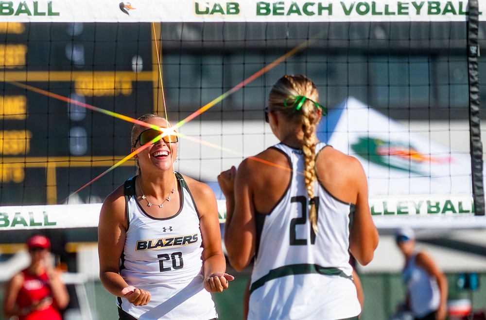 Two UAB volleyball players smile and congratulate each other in front of the net while a bright lens flare reflects of one player's sunglasses