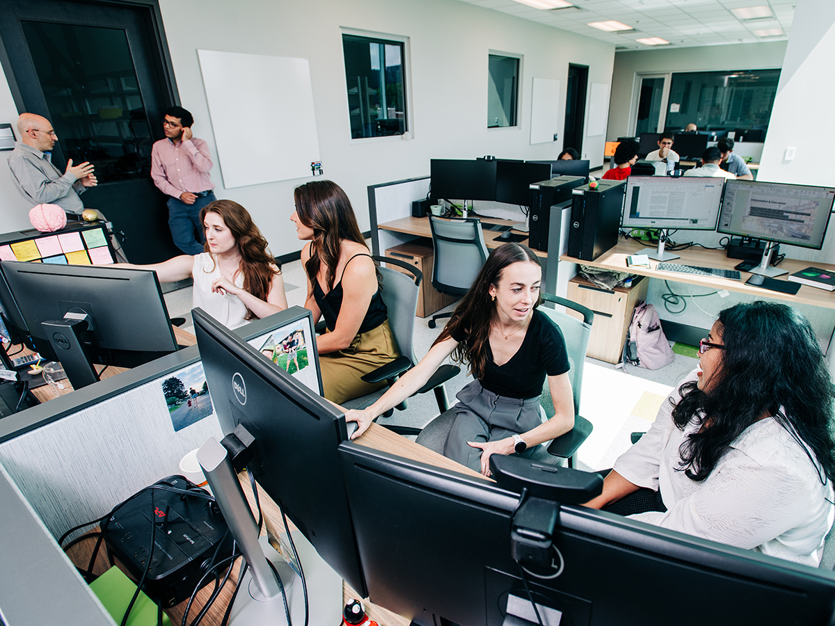 Four researchers in a UAB lab talk with each other in front of banks of computer monitors while other researchers and students occupy lab space behind them