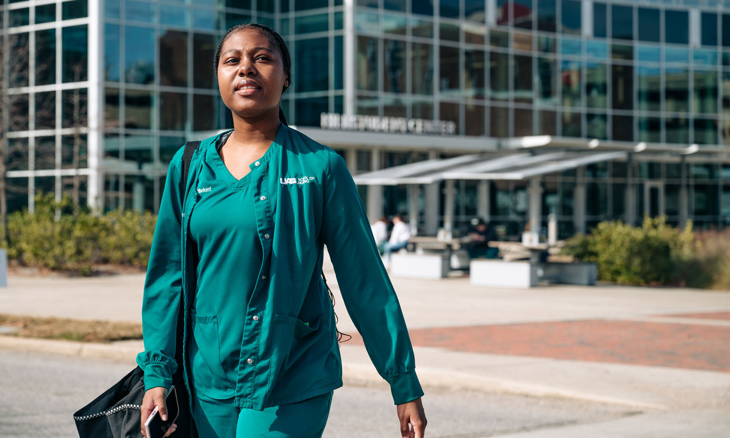 Black female nursing student wearing green scrubs walks across a crosswalk in front of UAB's Hill Student Center.