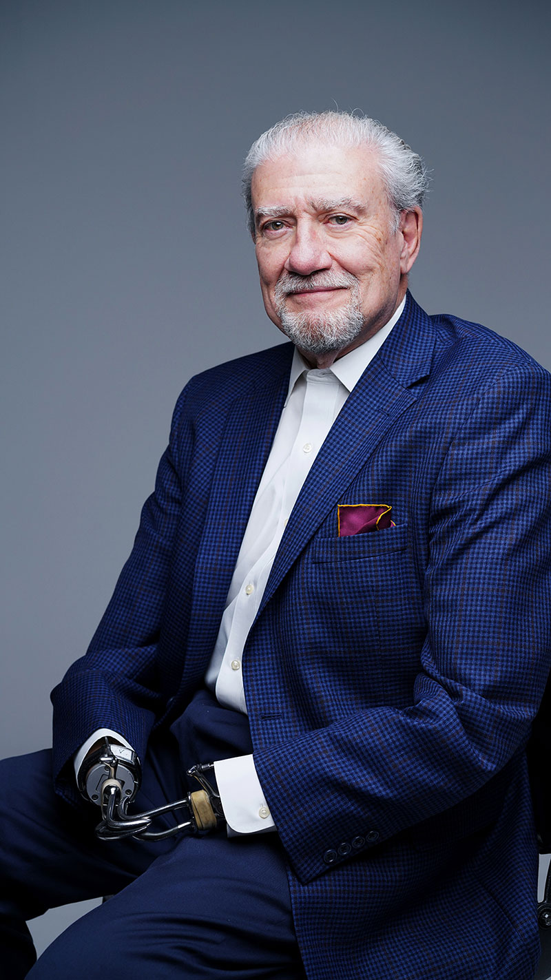 John Kemp sits with his prosthetics resting on his lap, wearing a stylish blue suit, maroon pocket square, and white collared shirt. 