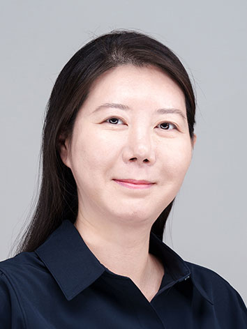 Dr. Kim has short dark hair. She's wearing a winder coat and a dark red shirt. A thin gold chain peaks out over the shirt's collar. 