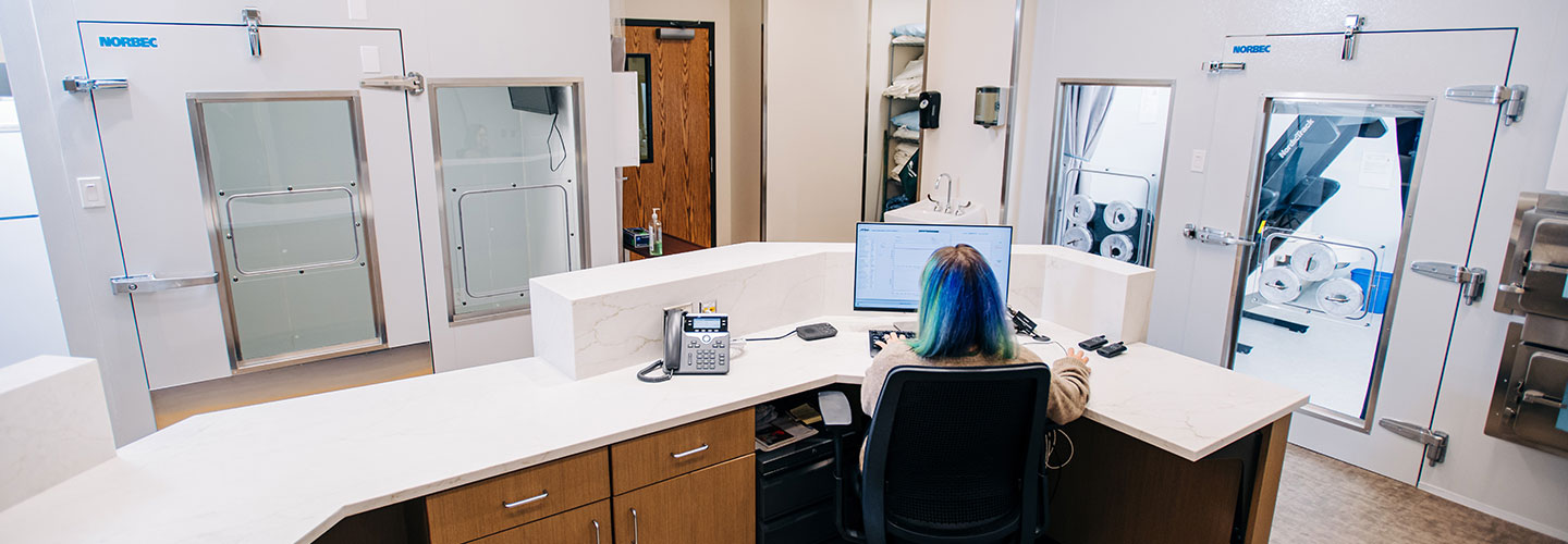 A person with shoulder-length blue/green hair works at the admin desk in the suite.
