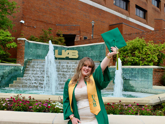 Wearing her graduation gown over beige clothes, Hutchinson lifts her graduation cap in front of a fountain.