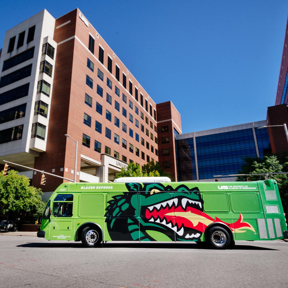 From side, a Blazer Express bus showing a graphic of Blaze (mascot) breathing fire traveling on 19th Street South in front of the North Pavilion of UAB Hospital.